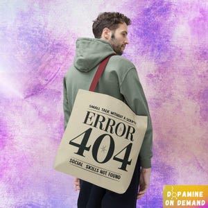 May include: A person wearing a green hoodie carries a beige tote bag with the text "Small talk without a script? Error 404 Social skills not found".