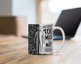 May include: White ceramic mug with a black and white graphic. The design features a split human figure and the words "SIMULTANEOUSLY TOO MUCH AND NOT ENOUGH" in bold, black text. The mug is on a wooden surface.
