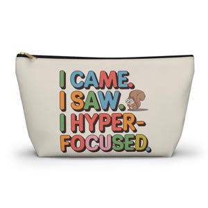 May include: A beige zippered pouch with the text "I CAME. I SAW. I HYPER-FOCUSED." in colorful letters. A cartoon squirrel is next to the text.