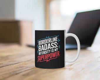 May include: A black ceramic coffee mug with the text "BORDERLINE BADASS: INTENSITY IS MY SUPERPOWER" in white, teal, and red. The mug has a white handle and is sitting on a wooden surface.