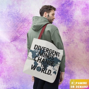 Könnte beinhalten: Eine weiße Einkaufstasche mit einem Weltkarten-Design und dem Text "Divergent Minds Change the World." Die Tasche hat rote Henkel und wird von einer Person getragen, die ein grünes Hoodie trägt.