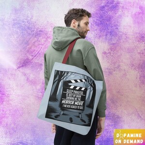 Może przedstawiać: Jasnoniebieska torba typu tote z czerwonym paskiem. Posiada wzór o tematyce horroru z klapsem filmowym w leśnym otoczeniu. Tekst na torbie brzmi: "SLEEP PARALYSIS IS JUST MY BRAIN SHOWING ME THE HORROR MOVIE I NEVER ASKED TO SEE."