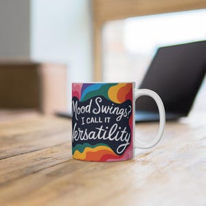Könnte beinhalten: Weiße Keramik-Tasse mit dem Text "Mood Swings? I call it Versatility" in einer verspielten Schriftart. Die Tasse hat ein farbenfrohes Regenbogen-Design. Die Tasse steht auf einer Holzoberfläche.