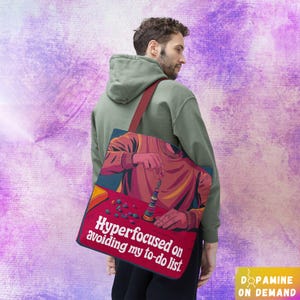 Op de afbeelding: Een rode en blauwe tote bag met een afbeelding van een persoon die een stapelspel speelt. De tekst op de tas luidt "Hyperfocused on avoiding my to-do list."