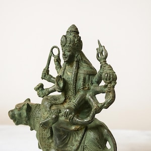 May include: A detailed bronze statue of a multi-armed deity, seated on a tiger. The figure wears a crown and ornate jewelry, holding various objects. The statue has a weathered, aged appearance, with a green patina.