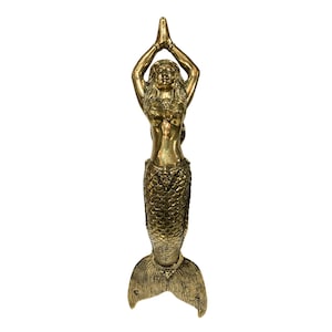 May include: A gold-colored mermaid statue with her hands raised in a prayer position. The mermaid has a detailed, scaly tail and a flowing, long hair.