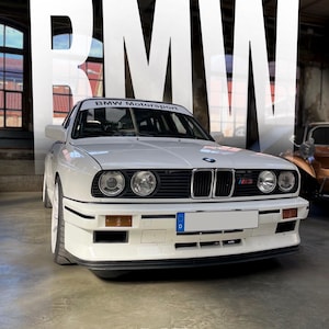 May include: A white BMW M3 car with the word "BMW" in large letters in the background. The car has a black front grill, headlights, and a "BMW Motorsport" banner on the windshield. The car is parked in a garage.