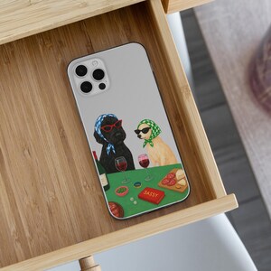 May include: A phone case featuring a whimsical illustration of two dogs at a table. One dog wears a blue headscarf and red glasses, the other a green headscarf and sunglasses. A bottle of wine, glasses, and a "SASSY" card are on the table.