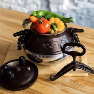 May include: A brown and silver pressure cooker with a lid and stand. The cooker is filled with fresh vegetables, including a tomato, yellow bell pepper, and green pepper. The cooker is on a wooden surface.