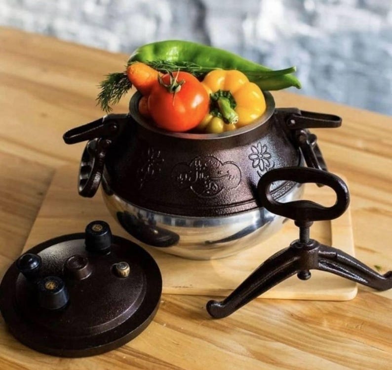 May include: A brown and silver pressure cooker with a lid and stand. The cooker is filled with fresh vegetables, including a tomato, yellow bell pepper, and green pepper. The cooker is on a wooden surface.