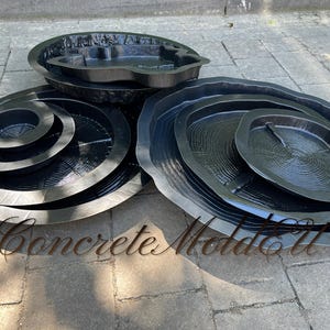 Puede incluir: Una colección de moldes de hormigón negro en varias formas y tamaños, incluyendo diseños circulares, ovalados y uno en forma de riñón. Los moldes tienen una superficie texturizada y están dispuestos sobre una superficie de piedra gris. El texto "Concrete Molds" es visible.