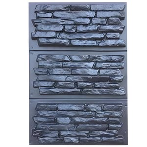 May include: A rectangular dark gray plastic mold featuring three stacked horizontal sections, each embossed with a detailed, irregular stone wall pattern. The surface displays varied stone shapes and textures designed for casting concrete or plaster.