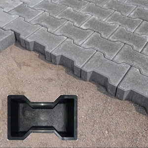 Puede incluir: Un molde de plástico negro para hacer adoquines de hormigón entrelazados. El molde tiene forma de flecha de doble punta. El molde está sentado sobre un lecho de arena. En el fondo, se puede ver un camino hecho de adoquines de hormigón entrelazados.