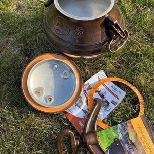 Puede incluir: Un kazan de metal marrón, una olla de cocina tradicional, con tapa y asa. La tapa está quitada, revelando el interior. La olla está sobre hierba, con componentes adicionales y materiales impresos cerca.