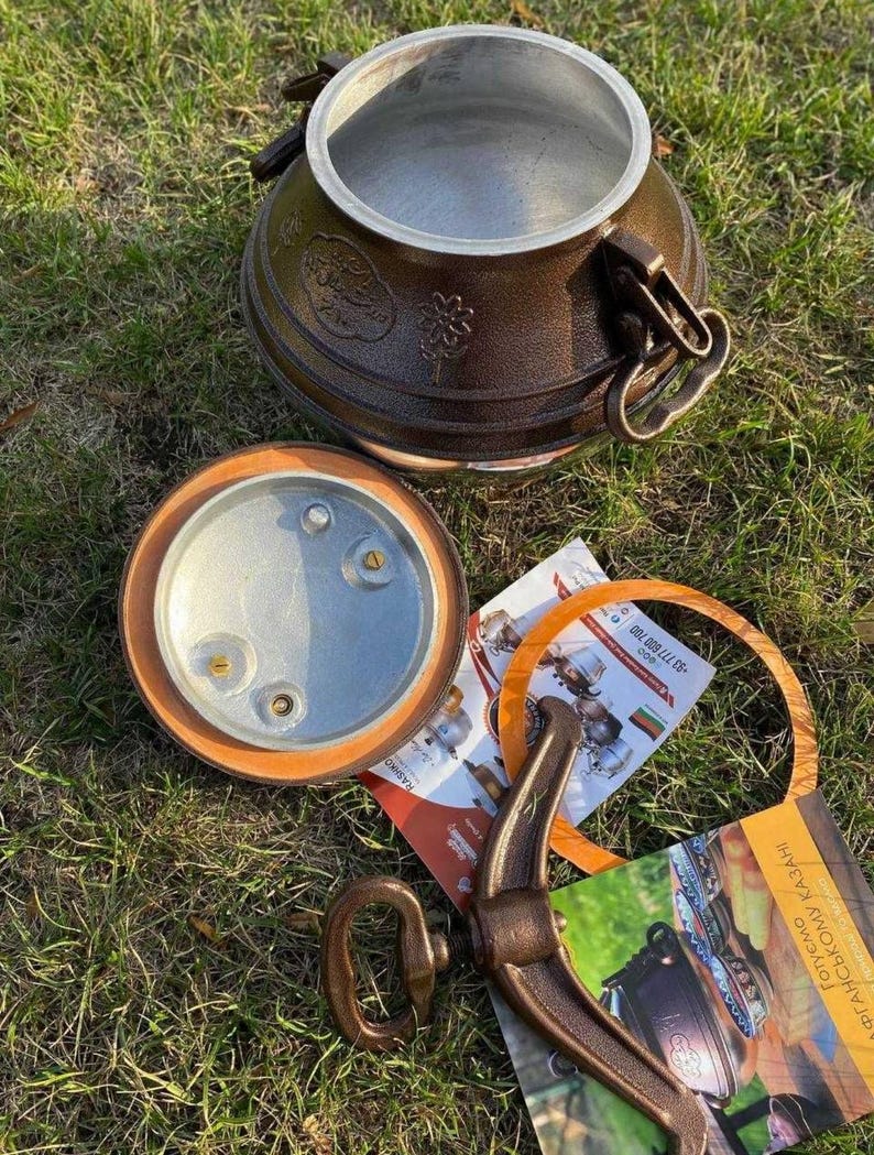 May include: A brown metal kazan, a traditional cooking pot, with a lid and handle. The lid is off, revealing the interior. The pot sits on grass, with additional components and printed materials nearby.
