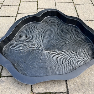 May include: A black, tree-ring-shaped tray with a textured surface. The tray has a dark gray border and a detailed, concentric circle pattern resembling tree rings. The tray is designed to resemble a cross-section of a tree trunk.