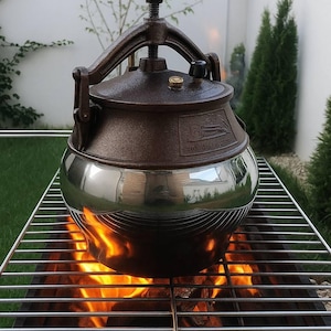 May include: A brown and silver pressure cooker on a metal grill above a fire. The cooker has a rounded base and a dark brown lid with a handle. Flames from the fire are visible beneath the grill, creating a warm, inviting scene.