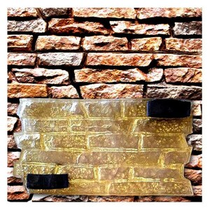 Può includere: Uno stampo rettangolare trasparente con un motivo a mattoni, con due cinturini in velcro neri. Lo stampo è posto su uno sfondo di un muro di mattoni naturali. Il motivo a mattoni è dettagliato e strutturato, con varie tonalità di marrone e beige.