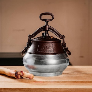 Puede incluir: Una olla a presión vintage con una base plateada cepillada y una parte superior y un asa de color marrón oscuro. El asa es arqueada y la parte superior tiene un diseño decorativo. La olla se encuentra sobre una superficie de madera.