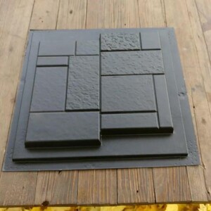 May include: A black paving stone mold with various rectangular and square shapes. The mold is set on a wooden surface, with yellow leaves scattered around the edges. The mold is designed for creating decorative concrete pavers.