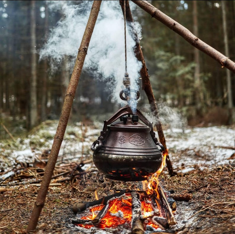 May include: A bronze-colored, antique-style cooking pot suspended over a campfire. The pot hangs from a tripod of wooden branches, with smoke rising from it. The fire below is bright orange and red, set in a forest setting.