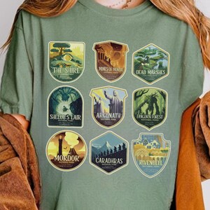 Op de afbeelding: Saliegroen T-shirt met negen geïllustreerde patches. De patches tonen scènes uit een fantasiewereld, waaronder "The Shire", "Mines of Moria" en "Mordor". Het shirt is een comfortabele, casual stijl.