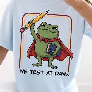 May include: Light blue t-shirt featuring a cartoon frog wearing a red cape and holding a pencil and a book. The text "WE TEST AT DAWN" is printed below the frog illustration. The shirt has a crew neck and short sleeves.