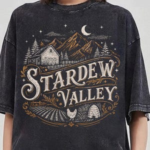 Puede incluir: Camiseta gris oscuro con un dise&ntilde;o vintage que dice "Stardew Valley". El dise&ntilde;o incluye un granero, monta&ntilde;as, &aacute;rboles, una luna creciente y un campo con una gallina y una colmena. La camiseta tiene un aspecto ligeramente descolorido.