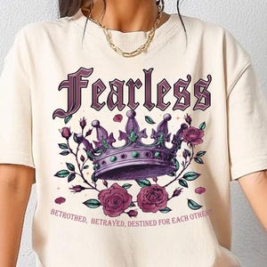May include: A cream colored t-shirt with a purple crown and pink roses. The text "Fearless" is printed in purple above the crown. The text "Betrothed, Betrayed, Destined for Each Other" is printed below the crown.