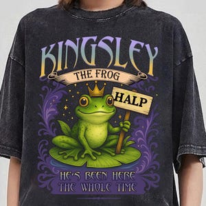 May include: Dark grey t-shirt with a cartoon frog wearing a crown and holding a sign saying "HALP." The shirt features the text "KINGSLEY THE FROG" above the frog and "HE'S BEEN HERE THE WHOLE TIME" below.