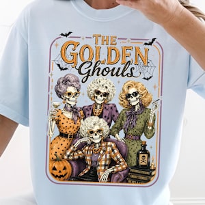 May include: A light blue t-shirt featuring a graphic print of four skeletons styled with blonde, grey, and auburn hair, wearing vintage clothing and sunglasses. The text reads THE GOLDEN Ghouls, accompanied by illustrations of a pumpkin, books, and bats.