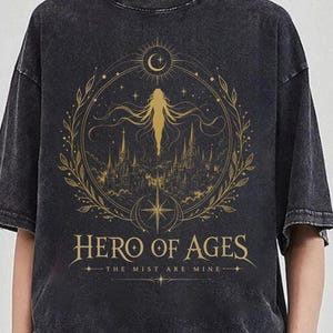 May include: Dark gray t-shirt with a gold illustration featuring a figure, a crescent moon, and a cityscape within a circular design. The text "HERO OF AGES" and "THE MIST ARE MINE" are printed below the illustration.