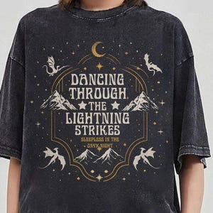 Könnte beinhalten: Dunkelgraues T-Shirt mit einem Grafikdesign. Der Text "Dancing Through The Lightning Strikes" ist von Illustrationen von Drachen, Bergen, einem Mond und Sternen umgeben. Das T-Shirt hat einen Vintage-Look.