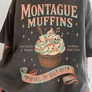 Op de afbeelding: Donkergrijs t-shirt met de tekst "MONTAGUE MUFFINS" en een cupcake-afbeelding met een schedel, botten en een mes. Extra tekst bevat "Best Muffins & Weapons" en "Providence Rhode Island". Een banner luidt "FRESH OUT THE BATCH OVEN".