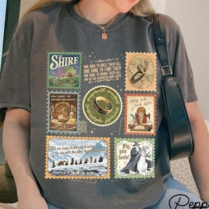 Op de afbeelding: Donkergrijs T-shirt met een collage van postzegelontwerpen met afbeeldingen en tekst uit een fantasieroman. De postzegels tonen scènes van een ring, personages en landschappen. Tekst bevat "Shire" en "Fly, you fools!"