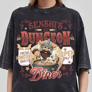 Könnte beinhalten: Schwarzes T-Shirt mit einem verwaschenen Grafikdesign. Das Design zeigt den Text "Senshi's Dungeon Diner" mit einer Illustration eines wikingerartigen Kochs, der in einem Topf rührt. Das Shirt enthält auch den Text "Fresh Monsters Daily" und "Open 24/7".