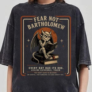 May include: A charcoal grey acid-wash t-shirt featuring a graphic of a gargoyle reading a book while sitting on a stack of books. The text reads FEAR NOT BARTHOLOMEW, EVERY DAY HAS ITS DOG, AISLING CATHEDRAL, TRAUM, and YOU DON'T HAVE TO BE GOOD.