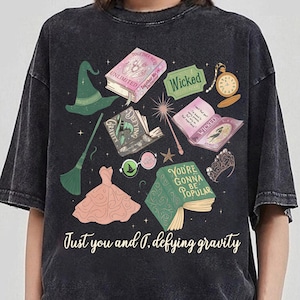 Könnte beinhalten: Dunkelgraues T-Shirt mit einem Fantasy-Motiv. Es zeigt Illustrationen von Büchern, einem Hexenhut, einem Kleid und einem Zauberstab. Der Text "Just you and I. defying gravity" ist darunter aufgedruckt. Ein lustiges Grafik-T-Shirt.