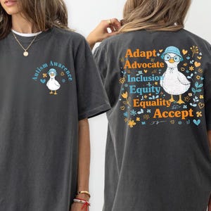May include: Dark gray t-shirt with a graphic of a duck wearing glasses and a hat, with the words "Autism Awareness" on the front. The back of the shirt features the words "Adapt, Advocate, Inclusion, Equity, Equality, Accept."