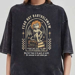 May include: A charcoal grey acid-wash t-shirt featuring a graphic of a gargoyle sitting on a stack of books. The text reads FEAR NOT BARTHOLOMEW, EVERY DAY HAS ITS DOG, AISLING CATHEDRAL, TRAUM, and You don't have to be good, or useful, for someone to care about you.