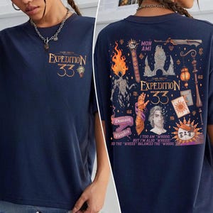 Puede incluir: Camiseta azul marino con texto dorado "EXPEDITION 33" en la parte delantera. La parte trasera presenta un gráfico detallado con texto que incluye "MON AMI", "Demain Viendra" y la frase "I TOO AM 'WHOOO' BUT I'M ALSO 'WHEEE!'".