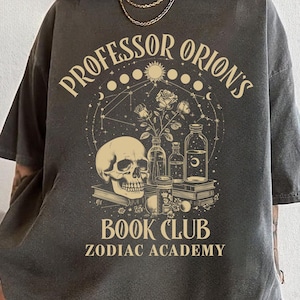 Puede incluir: Una camiseta gris oscuro con un dise&ntilde;o gr&aacute;fico que presenta una calavera, rosas y botellas. El texto "Professor Orion's Book Club Zodiac Academy" est&aacute; impreso en la camiseta.