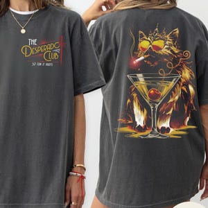 May include: Pepper-colored t-shirt with a graphic on the front and back. The front features text that reads "The Desperado Club, So Fun It Hurts." The back displays a cat wearing sunglasses and a crown, smoking a cigarette, and next to a martini glass.