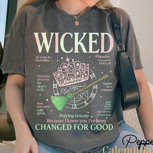 Könnte beinhalten: Dunkelgraues T-Shirt mit dem Wort "WICKED" in Rosa und Weiß. Das Shirt zeigt eine rosa Krone, einen grünen Besen und Text aus dem Musical. Das Shirt hat auch den Text "Defying Gravity" und "Changed for Good".