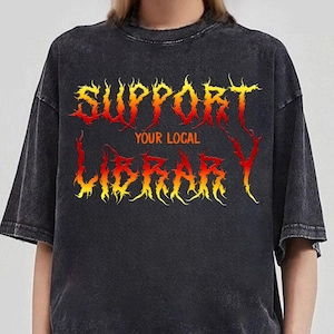 May include: Dark gray t-shirt with the text "SUPPORT YOUR LOCAL LIBRARY" in a fiery, yellow and red font. The shirt has a vintage, washed look and a crew neck.
