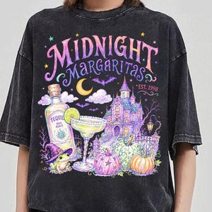 May include: A charcoal grey acid-wash t-shirt featuring a colorful graphic print with the text "MIDNIGHT MARGARITAS" and "EST. 1998". The design includes a tequila bottle, a margarita glass, a haunted house, pumpkins, and a frog in a witch hat.