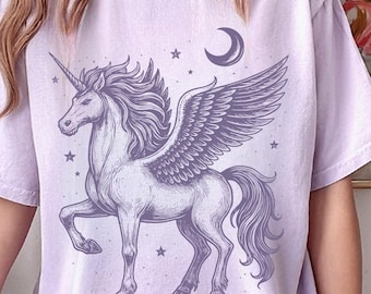 May include: A light purple t-shirt featuring a detailed illustration of a winged unicorn, a crescent moon, and stars. The unicorn is depicted in shades of purple and gray. The text "CalendulaStyle" is at the bottom right.