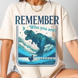 Puede incluir: Camiseta color crema con un diseño gráfico de un Tyrannosaurus Rex turquesa y el texto "REMEMBER Who you are!". El diseño incluye un paisaje azul y blanco con una gallina, enmarcado en un rectángulo azul oscuro.