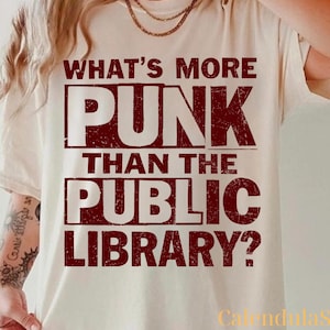 以下が含まれることがあります： クリーム色のTシャツ。テキスト「WHAT'S MORE PUNK THAN THE PUBLIC LIBRARY?」は、使い古されたダークレッドのフォントで表示されています。このシャツは半袖で、クルーネックです。