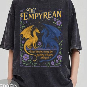 Może przedstawiać: Ciemnoszara koszulka z nadrukiem z napisem "The Empyrean" nad ilustracją złotego i niebieskiego smoka. Poniżej smok&oacute;w znajduje się fraza "I had the time of my life fighting dragons with you". Fioletowe kwiaty otaczają wz&oacute;r.